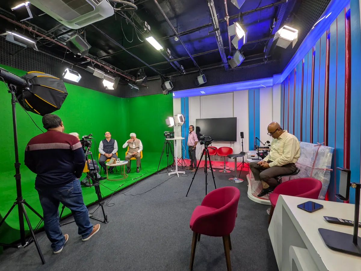A television studio mid-shoot: lighting rigs, a green screen, a camera operator and crew working around an interview in progress.